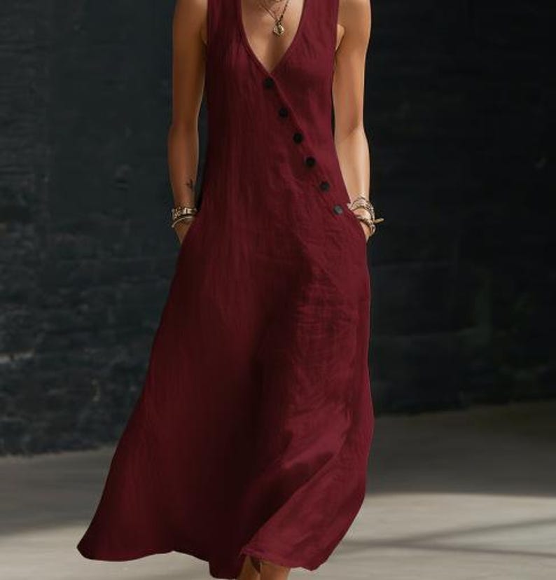 May include: A sleeveless, burgundy maxi dress with a V-neckline and a button-up closure. The dress features two pockets and a flowing, floor-length skirt. The fabric appears to be a lightweight linen or cotton blend, suitable for warm weather.