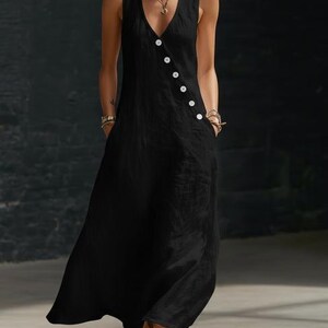 May include: A sleeveless black dress with a V-neckline and a row of white buttons down the side. The dress has pockets and a flowing, floor-length skirt. The fabric appears to be a lightweight material, suitable for warm weather.