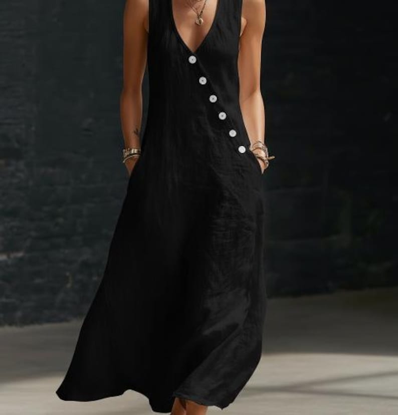 May include: A sleeveless black dress with a V-neckline and a row of white buttons down the side. The dress has pockets and a flowing, floor-length skirt. The fabric appears to be a lightweight material, suitable for warm weather.