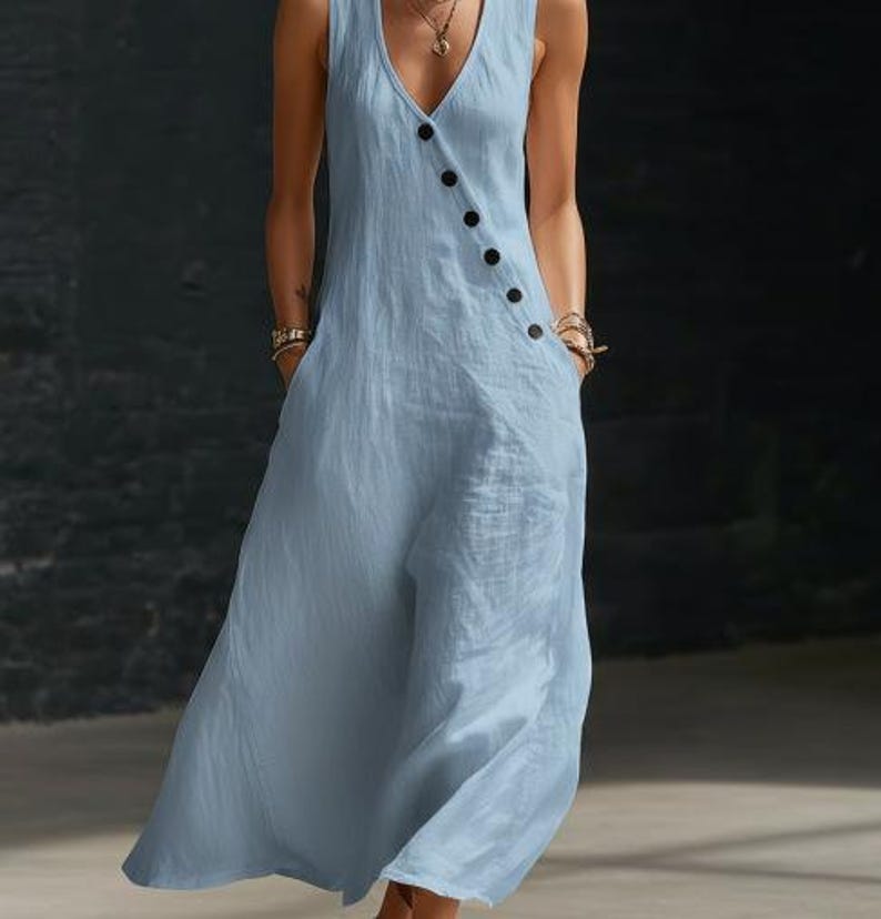 May include: A sleeveless, light blue jumpsuit with a V-neckline and a row of black buttons down the side. The jumpsuit has pockets and a flowing, wide-leg design, made of a soft, textured fabric.
