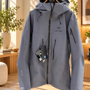 Könnte beinhalten: Eine schieferblaue Arc'teryx Kapuzenjacke mit durchgehendem schwarzem Frontreißverschluss, zwei vertikalen Brusttaschen und einer Ärmeltasche. Die Jacke hat ein Gore-Tex-Logo am Ärmelbündchen sowie angehängte Produktetiketten mit XL und Arc'teryx.