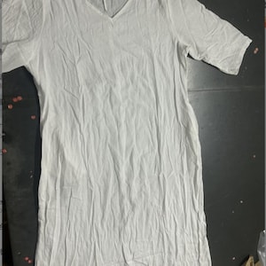 May include: A white, V-neck dress with short sleeves. The dress is made of a lightweight, textured fabric, and has a relaxed fit. The garment is laid flat on a dark surface.