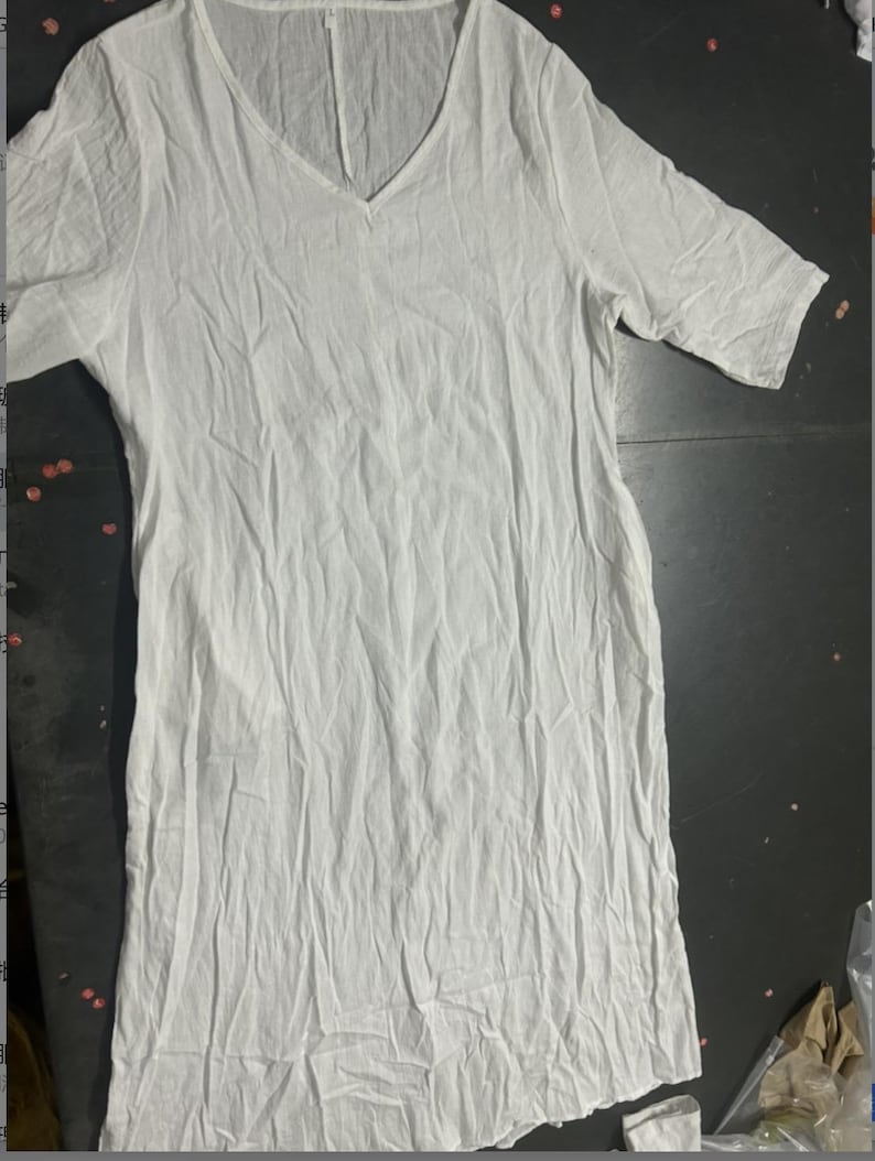 May include: A white, V-neck dress with short sleeves. The dress is made of a lightweight, textured fabric, and has a relaxed fit. The garment is laid flat on a dark surface.