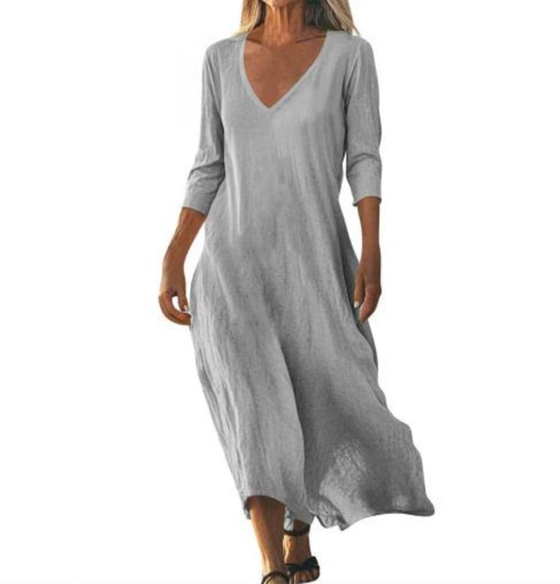 May include: A light gray, V-neck dress with three-quarter sleeves. The dress has a loose, flowing design and falls to the mid-calf. The fabric appears to be a lightweight linen or cotton blend, suitable for warm weather.