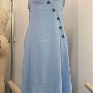May include: A light blue sleeveless dress with a V-neckline and a series of dark buttons down the side. The dress is displayed on a mannequin, showcasing its flowing, casual design. The dress has a relaxed fit and a midi length.