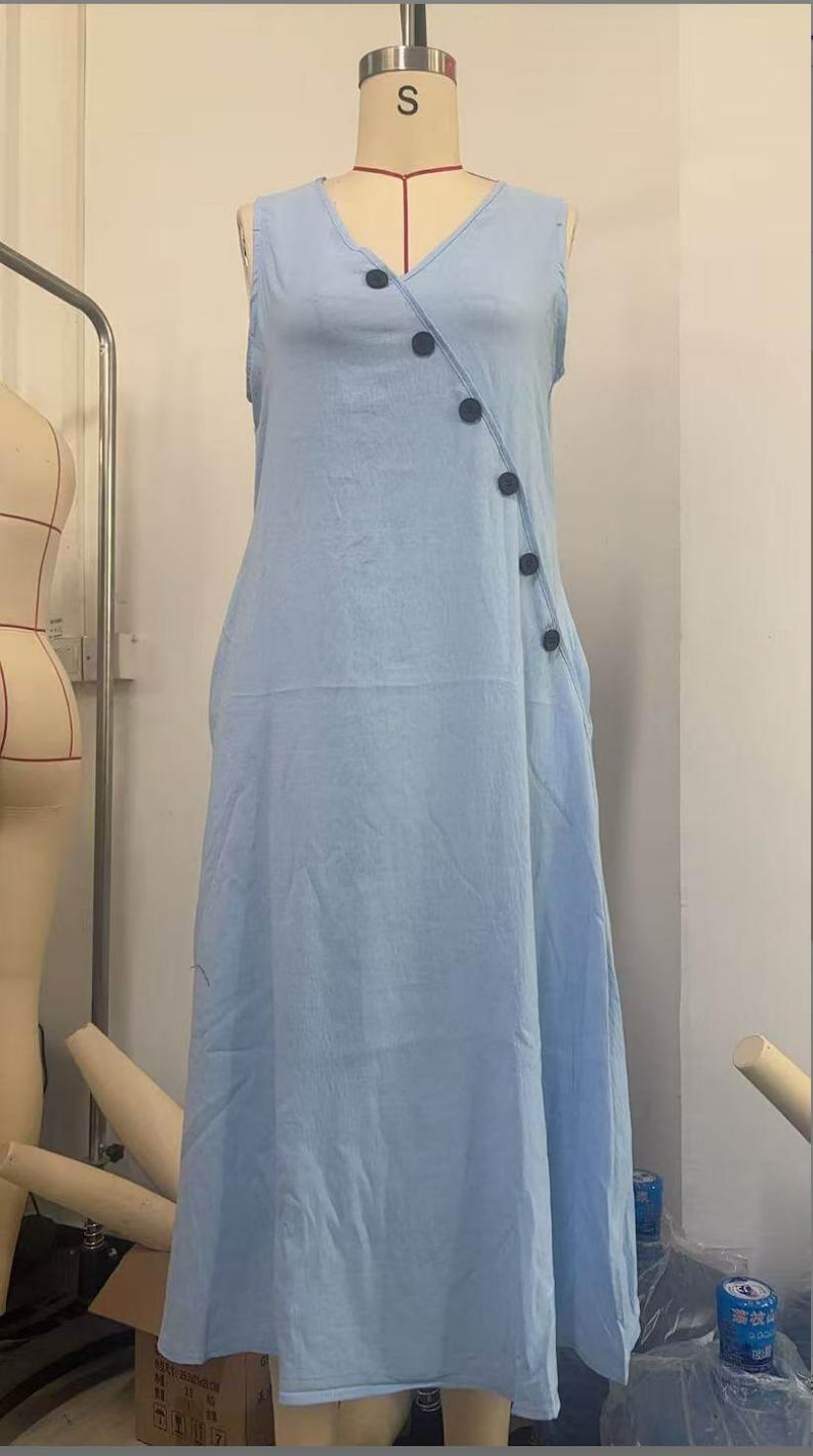 May include: A light blue sleeveless dress with a V-neckline and a series of dark buttons down the side. The dress is displayed on a mannequin, showcasing its flowing, casual design. The dress has a relaxed fit and a midi length.