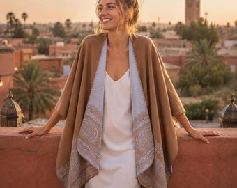 May include: A tan and light blue reversible open-front wrap featuring a detailed paisley pattern along the edges. The garment is worn over a simple white slip dress, showcasing a soft texture and a draped silhouette that falls loosely over the shoulders.
