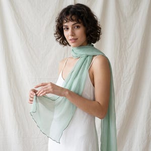 May include: A light sage green scarf with a delicate, sheer texture and a finished edge, draped around the neck and shoulders over a simple white sleeveless garment. The fabric is lightweight and semi-transparent, showing soft folds.