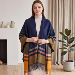 Puede incluir: Un poncho abierto de cuadros en azul marino, amarillo mostaza y naranja quemado con textura de espiga y flecos en el dobladillo inferior. La prenda se lleva sobre un suéter de cuello alto beige y presenta una silueta amplia y drapeada.