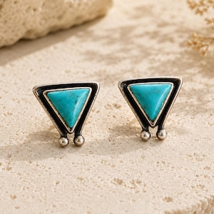 Puede incluir: Un par de pendientes de botón con una piedra turquesa triangular engastada en un marco de metal plateado. El diseño incluye una parte superior puntiaguda y dos pequeños acentos esféricos en la base de cada pendiente, creando un aspecto geométrico.