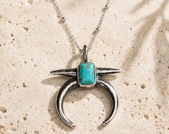 May include: A silver-toned necklace featuring a crescent-shaped pendant with a hammered texture and a rectangular turquoise stone set in the center. The pendant hangs from a delicate silver chain with small, evenly spaced spherical beads.