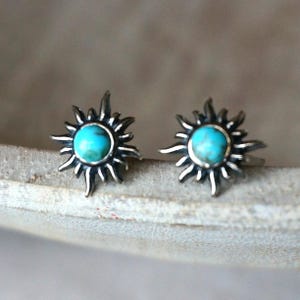 May include: A pair of silver stud earrings. Each earring has a turquoise stone at the centre, encircled by a sunburst design. The earrings are displayed against a neutral background.