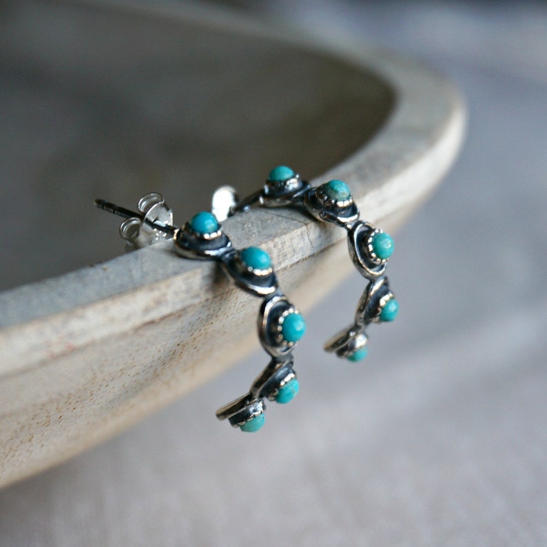 May include: Silver hoop earrings with turquoise stones. The earrings are a half-circle shape and have a total of seven turquoise stones.