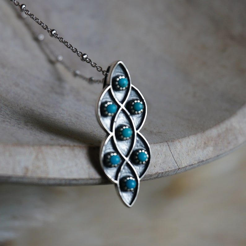 Puede incluir: Un collar de plata con un colgante de dise&ntilde;o geom&eacute;trico con piedras turquesa. El colgante tiene forma de l&aacute;grima y presenta un patr&oacute;n repetitivo de c&iacute;rculos entrelazados.