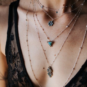 Puede incluir: Primer plano de una persona que lleva varias cadenas de plata con piedras turquesa. Las cadenas presentan una variedad de estilos de colgantes, incluyendo una flor, un tri&aacute;ngulo y un grupo de piedras.