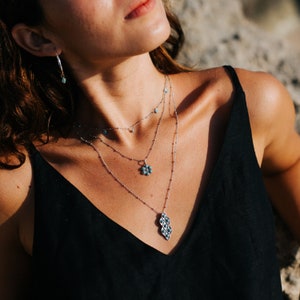 Puede incluir: Una mujer lleva un top negro de tirantes con tres collares de plata con piedras turquesa. Los collares tienen un colgante de flor y un colgante geom&eacute;trico.