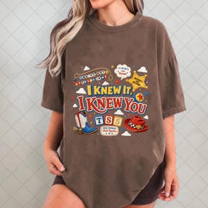 May include: A brown short-sleeve t-shirt featuring a graphic print with the text "I KNEW IT, I KNEW YOU" and "TS5". The design includes a cowboy boot, a red cowboy hat, a yellow sheriff star, and various colorful decorative elements.