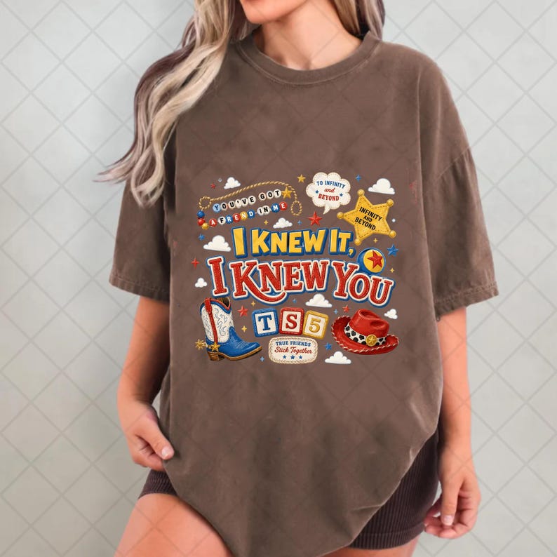 May include: A brown short-sleeve t-shirt featuring a graphic print with the text "I KNEW IT, I KNEW YOU" and "TS5". The design includes a cowboy boot, a red cowboy hat, a yellow sheriff star, and various colorful decorative elements.