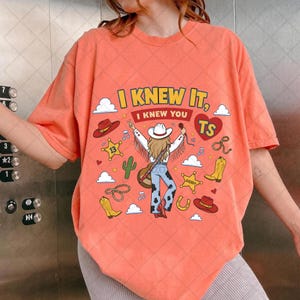 May include: An orange t-shirt featuring a graphic print of a person in a cowboy hat and fringe jacket, blue cow-print pants, and boots. The design includes the text I KNEW IT, I KNEW YOU TS, along with cowboy hats, stars, boots, and cacti.