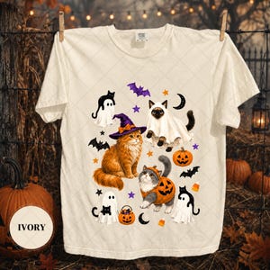 May include: An ivory t-shirt featuring a graphic print of three cats in Halloween costumes, including a ginger cat in a witch hat, a cat in a ghost sheet, and a cat in a pumpkin suit, surrounded by bats, stars, moons, and candy corn icons.
