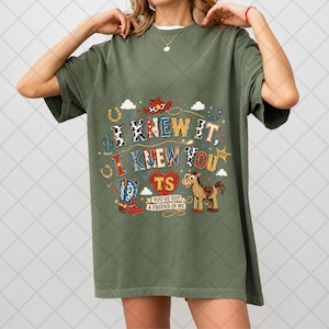 May include: An olive green t-shirt featuring a graphic print with the text I KNEW IT, I KNEW YOU, TS, and YOU'VE GOT A FRIEND IN ME. The design includes a cowboy hat, a blue and red cowboy boot, a brown horse, horseshoes, and a sheriff star.