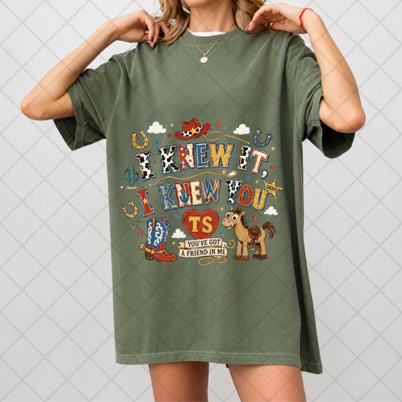 May include: An olive green t-shirt featuring a graphic print with the text I KNEW IT, I KNEW YOU, TS, and YOU'VE GOT A FRIEND IN ME. The design includes a cowboy hat, a blue and red cowboy boot, a brown horse, horseshoes, and a sheriff star.
