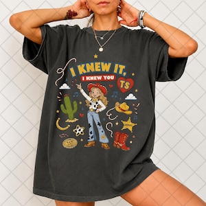 May include: A charcoal gray t-shirt featuring a graphic print of a cowgirl character, a cactus, a sheriff star, a cowboy hat, and red boots. The text reads I KNEW IT, I KNEW YOU TS, with a red heart icon. The design includes various western-themed elements.