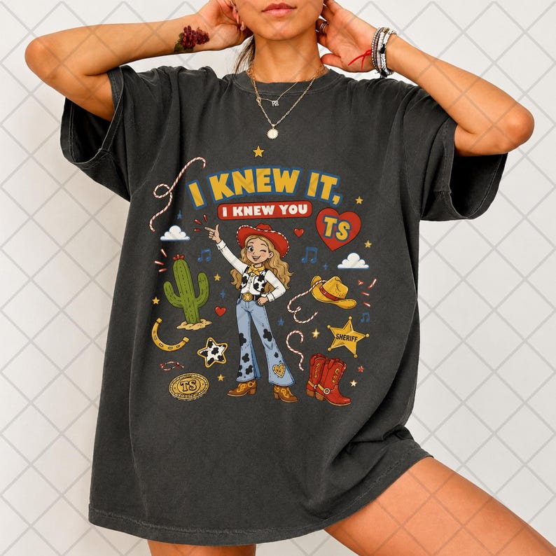 May include: A charcoal gray t-shirt featuring a graphic print of a cowgirl character, a cactus, a sheriff star, a cowboy hat, and red boots. The text reads I KNEW IT, I KNEW YOU TS, with a red heart icon. The design includes various western-themed elements.