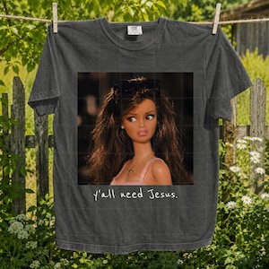 May include: A charcoal grey short-sleeved t-shirt featuring a graphic print of a doll with long brown hair, sunglasses on her head, and a gold cross necklace. Below the doll, the text reads, y'all need Jesus. The shirt has a crew neckline.