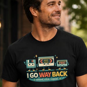Może przedstawiać: Czarny t-shirt z grafiką przedstawiającą dyskietkę, kasetę VHS i kasetę magnetofonową z uśmiechniętymi buźkami. Pod postaciami znajduje się napis I GO WAY BACK oraz rewind, click, repeat. Wzór wykorzystuje kolory niebieski, pomarańczowy i biały.