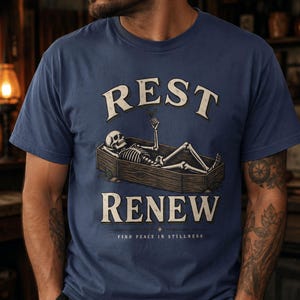 May include: A navy blue t-shirt featuring a graphic of a skeleton lying in a wooden coffin. The text "REST" appears above the coffin, "RENEW" below it, and "FIND PEACE IN STILLNESS" at the bottom. The skeleton holds a small butterfly.