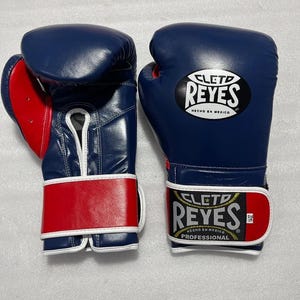 Könnte beinhalten: Ein Paar blaue und rote Boxhandschuhe mit dem Cleto Reyes Logo auf dem Handgelenkband und dem Handrücken. Die Handschuhe haben weiße Ränder, eine Schnürung an der Handfläche und den Schriftzug Cleto Reyes Hecho en Mexico Professional.