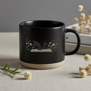 May include: Black ceramic mug with a dragon illustration. The mug features a black dragon on an open book, surrounded by white flowers. The bottom of the mug is a light beige color. The mug has a curved handle.