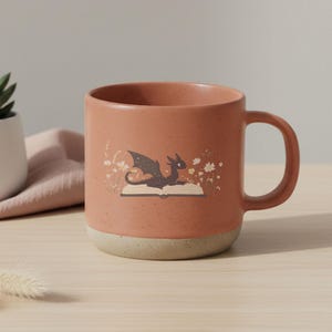 May include: A speckled terracotta-colored ceramic mug with a light beige base. The mug features a whimsical illustration of a dragon resting on an open book, surrounded by small white flowers. The handle is a matching terracotta color.