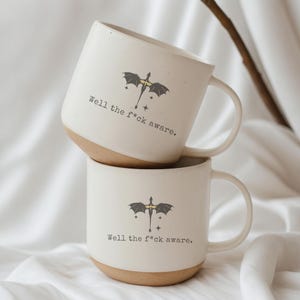 May include: Two white ceramic mugs with a tan base are stacked. Each mug features a black dragon graphic with a yellow center and the text "Well the f*ck aware." The mugs have a curved handle.