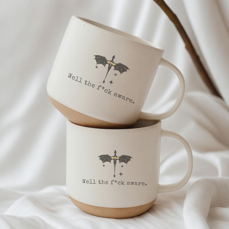 May include: Two white ceramic mugs with a tan base are stacked. Each mug features a black dragon graphic with a yellow center and the text "Well the f*ck aware." The mugs have a curved handle.