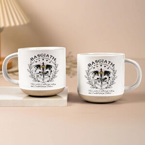 May include: Two white ceramic mugs with the text "BASGIATH WAR COLLEGE" and a graphic design. The mugs have a beige base and a curved handle. The design includes a sword, wings, and a quote.