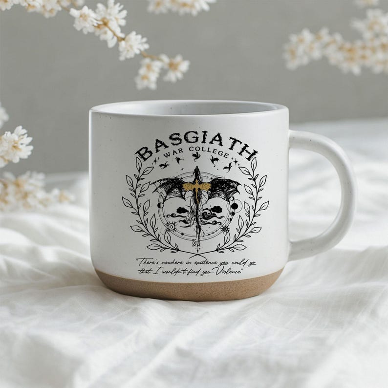 May include: White ceramic mug with a brown base, featuring the text "BASGIATH WAR COLLEGE" and a detailed graphic design. The design includes a sword, wings, and the quote, "There's nowhere in existence you could go that I wouldn't find you. Violence."