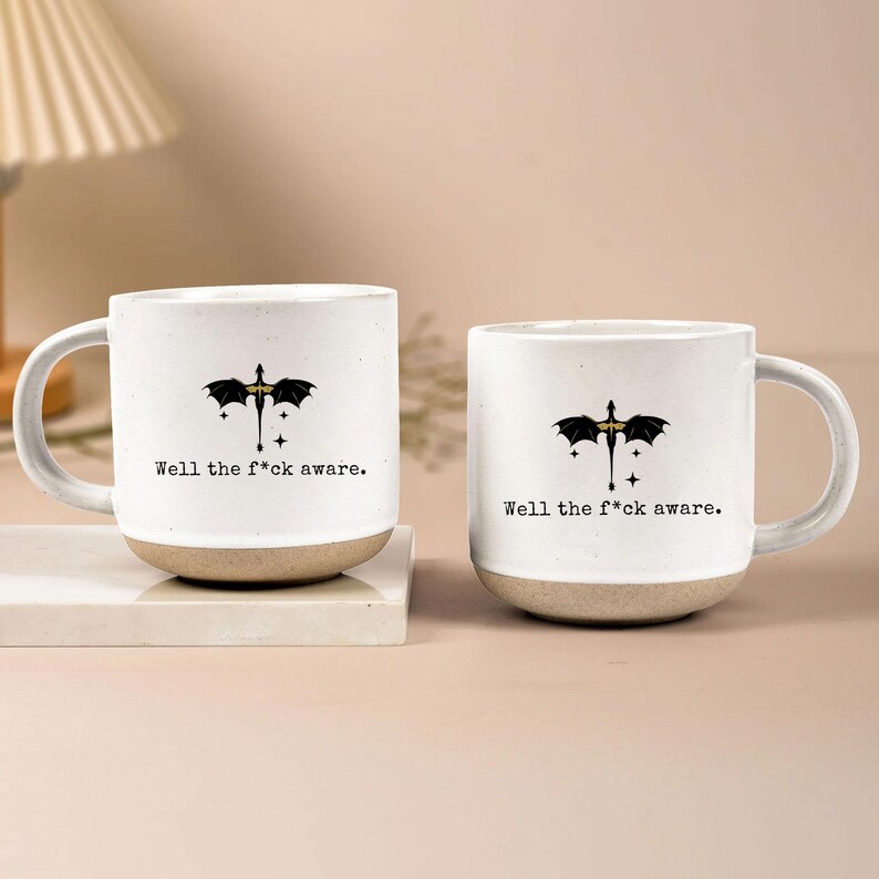 May include: Two white ceramic mugs with a dragon graphic and the text "Well the f*ck aware." The mugs have a beige base and a curved handle. The dragon graphic is black and gold.