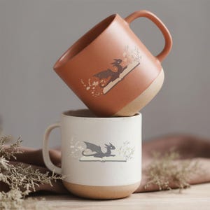 May include: Two ceramic mugs, one stacked on top of the other. The top mug is a speckled terracotta color, and the bottom mug is white. Each mug features a dragon illustration on an open book with floral accents.