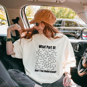 Pode incluir: Uma t-shirt de manga curta em tom off-white com uma impressão gráfica preta de várias estruturas moleculares químicas. O texto "What Part Of" está impresso acima das estruturas e "Don't You Understand?" abaixo do gráfico.