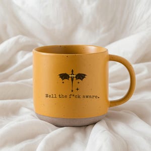 May include: Mustard yellow ceramic mug with a dragon graphic and the text "Well the f*ck aware." The mug has a speckled finish and a gray base. The handle is the same color as the mug.