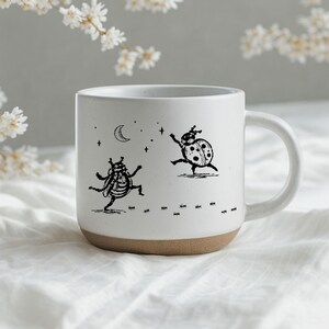 Peut inclure: Un mug en céramique blanche avec une finition mouchetée et une base en argile naturelle. L'avant présente une illustration à l'encre noire de deux coccinelles dansant sous un croissant de lune et des étoiles, avec une ligne de fourmis.