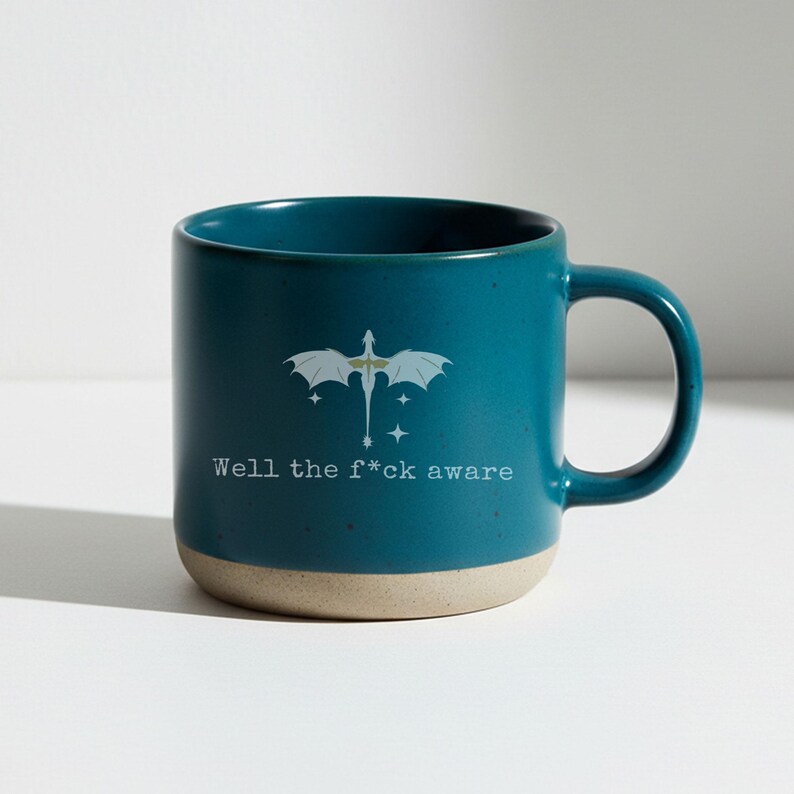 May include: Teal ceramic mug with a beige base and a curved handle. The mug features a white dragon graphic and the text "Well the f*ck aware". The mug is designed for hot beverages.
