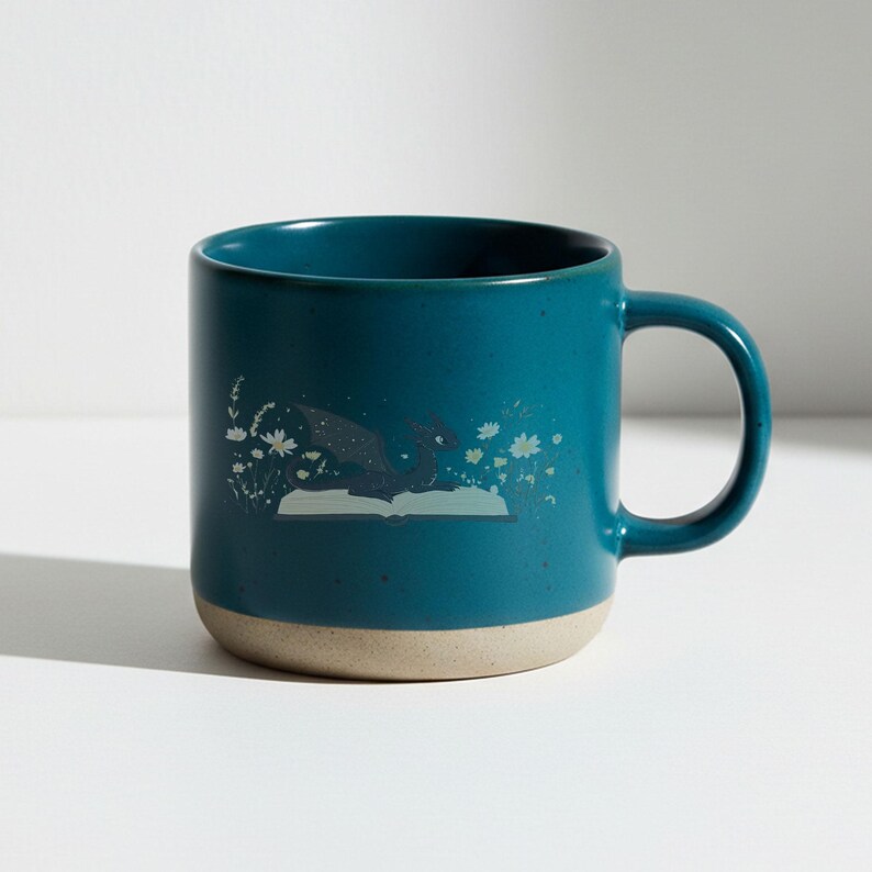May include: Teal ceramic mug with a beige base and a curved handle. The mug features a whimsical illustration of a dragon emerging from an open book, surrounded by floral and star accents. The mug is designed for hot beverages.