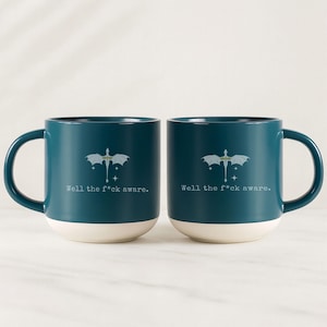 May include: Two teal ceramic mugs with white bottoms and large handles. Each mug features a dragon graphic and the text "Well the f*ck aware." The mugs are identical and sit side-by-side.