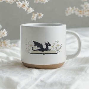 May include: A white ceramic mug with a brown base. The mug features a black dragon illustration sitting on an open book, surrounded by white and yellow flowers. The mug has a curved handle.