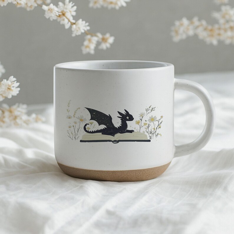 May include: A white ceramic mug with a brown base. The mug features a black dragon illustration sitting on an open book, surrounded by white and yellow flowers. The mug has a curved handle.
