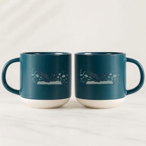 May include: Two teal ceramic mugs with white bottoms and large handles. Each mug features a whimsical illustration of a dark shape over an open book with floral accents. The mugs are set against a white background.