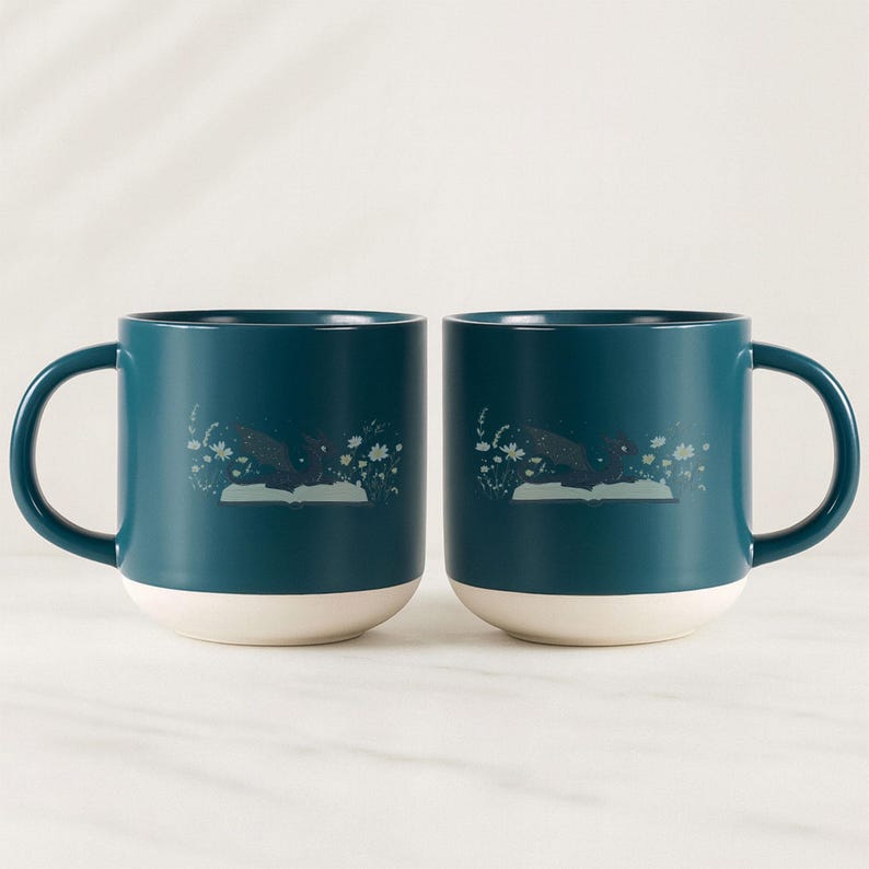 May include: Two teal ceramic mugs with white bottoms and large handles. Each mug features a whimsical illustration of a dark shape over an open book with floral accents. The mugs are set against a white background.